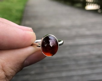 Könnte beinhalten: Ein silberner Ring mit einem dunkelroten ovalen Edelstein.