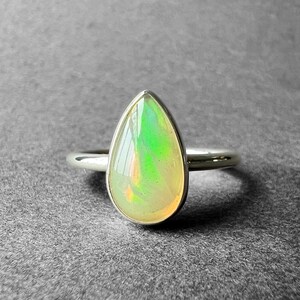 Può includere: Un anello con un opale a forma di lacrima incastonato in argento sterling. L'opale è di colore verde pallido e bianco con una lucentezza madreperlacea.