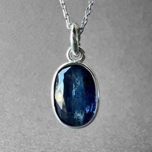 May include: A silver necklace featuring an oval-shaped, faceted deep blue kyanite gemstone pendant. The stone is set in a simple silver bezel frame and hangs from a delicate silver chain, showcasing natural inclusions and a polished surface.
