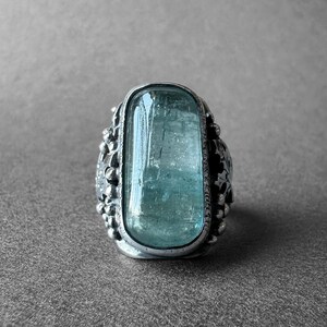 Op de afbeelding: Een zilveren ring met een grote, rechthoekige, lichtblauwe aquamarijn, gezet in een gedetailleerde zilveren zetting.