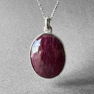 May include: A silver necklace with a large, oval-shaped, faceted ruby pendant. The ruby is a deep, rich red color and is set in a silver bezel. The chain is delicate and hangs from the pendant.