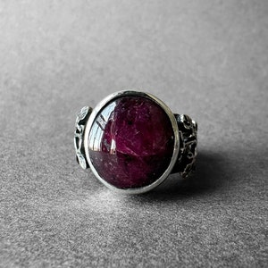 Peut inclure: Une bague en argent avec une grande pierre précieuse ronde de couleur rouge foncé sertie dans un sertissage à bélière. La bande de la bague est décorée de motifs floraux complexes.