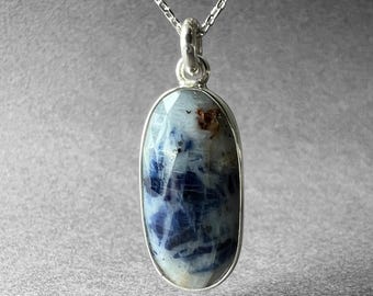 May include: A silver pendant necklace featuring an oval-shaped sodalite stone. The stone has a mix of blue, white, and brown colors. The pendant is encased in a silver setting and hangs from a delicate silver chain.