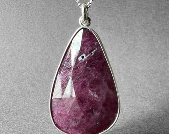 Könnte beinhalten: Halskette mit einem tropfenförmigen Anhänger aus Silber, besetzt mit einem großen, facettierten Rubin. Der Rubin hat eine tiefe rosa Farbe und ist in eine silberne Fassung gefasst. Der Anhänger hängt an einer feinen Silberkette.