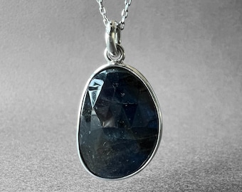 May include: A necklace featuring a faceted, dark blue, oval-shaped gemstone pendant set in a polished silver-toned metal bezel. The pendant hangs from a delicate silver-toned metal chain with small, uniform links, creating a simple and elegant jewelry piece.