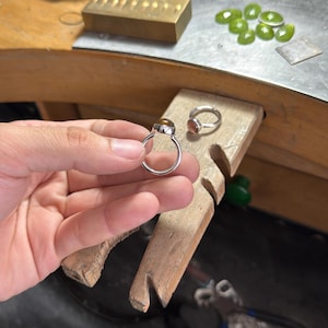 May include: Two silver rings featuring round, polished brown gemstones set in simple metal bands. One ring is held up, showcasing the smooth, domed surface of the stone, while the second ring rests on a wooden workbench surface nearby.