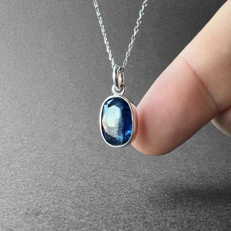 May include: A silver necklace featuring an oval-shaped, faceted deep blue gemstone pendant. The stone is set in a polished silver bezel and hangs from a delicate silver chain, showcasing a reflective surface with dark and light blue tones.