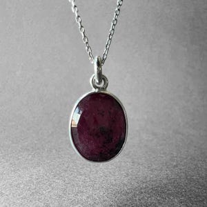 May include: A silver necklace featuring an oval-shaped pendant with a deep red, faceted gemstone. The pendant is suspended from a delicate silver chain, showcasing a polished metal setting that secures the dark, textured stone in place.