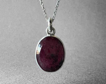 May include: A silver necklace featuring an oval-shaped pendant with a deep red, faceted gemstone. The pendant is suspended from a delicate silver chain, showcasing a polished metal setting that secures the dark, textured stone in place.