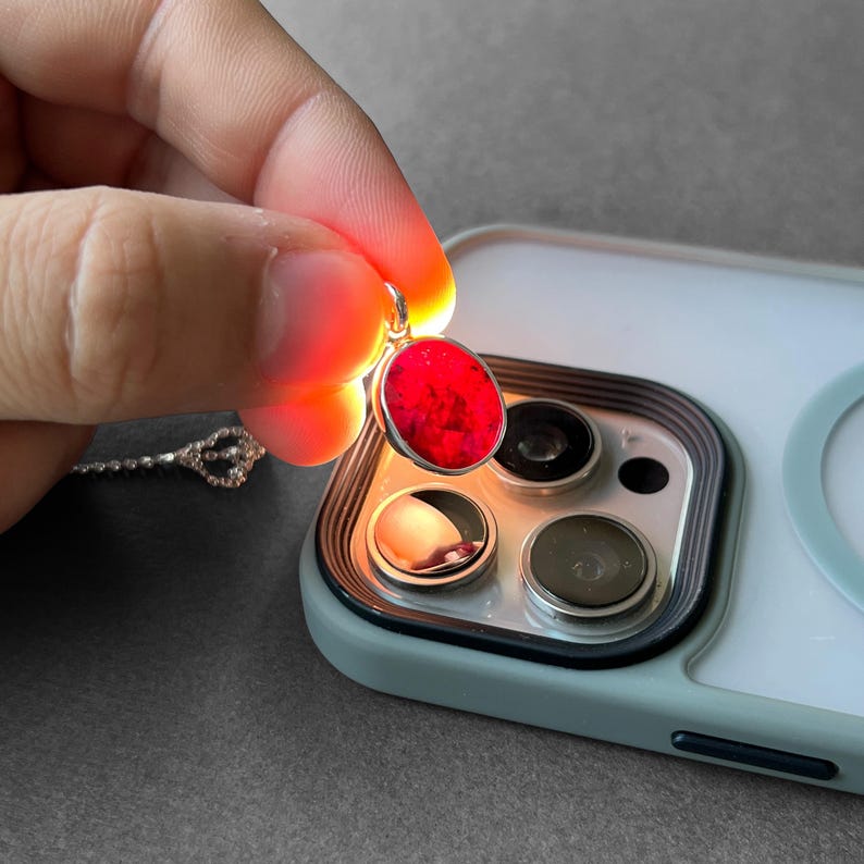 Op de afbeelding: Een ovale hanger met een levendige rode steen in een zilverkleurige metalen zetting. De hanger wordt boven de cameralenzen van een smartphone gehouden, waardoor de steen van achteren wordt verlicht en de dieprode kleur wordt benadrukt.