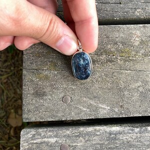 Peut inclure: Un pendentif en argent avec une pierre pr&eacute;cieuse ovale bleu fonc&eacute;. Le pendentif est tenu dans une main contre un fond en bois.