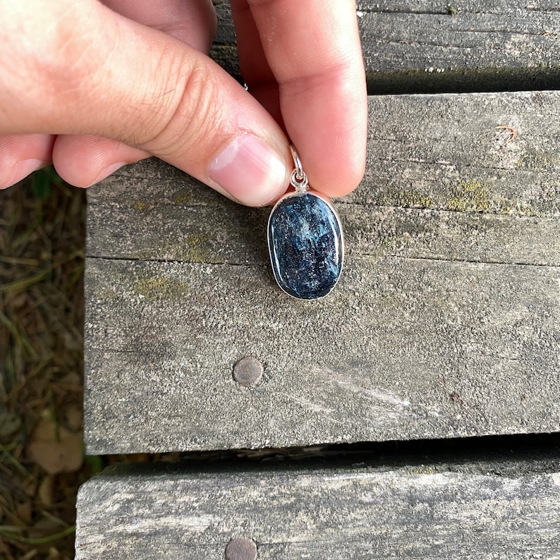 Peut inclure: Un pendentif en argent avec une pierre pr&eacute;cieuse ovale bleu fonc&eacute;. Le pendentif est tenu dans une main contre un fond en bois.