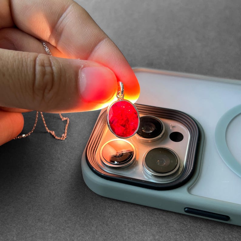 Op de afbeelding: Een ovaalvormige hanger met een levendige rode edelsteen in een zilverkleurige metalen zetting. De hanger hangt aan een fijne zilveren ketting en wordt van achteren verlicht, waardoor de rode steen intens oplicht tegen het licht.