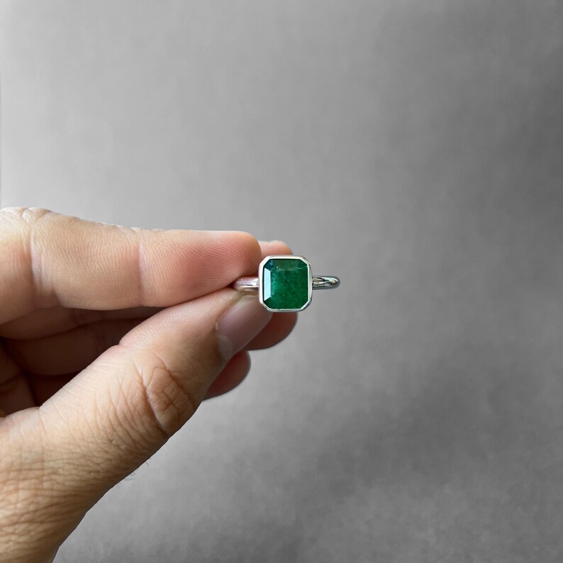K&ouml;nnte beinhalten: Ein Silberring mit einem rechteckigen, smaragdgeschliffenen gr&uuml;nen Edelstein in einer glatten Zargenfassung. Der Ring verf&uuml;gt &uuml;ber eine polierte Silberschiene, die mit dem Metallrahmen verbunden ist, der den leuchtend gr&uuml;nen Stein umschlie&szlig;t.