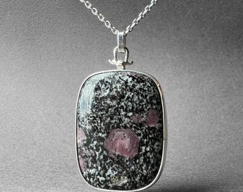 Könnte beinhalten: Eine silberne Kettenkette mit einem Anhänger, der einen großen, rechteckigen, schwarz-weißen Stein mit rosa Akzenten zeigt. Der Stein hat ein gesprenkeltes Muster.