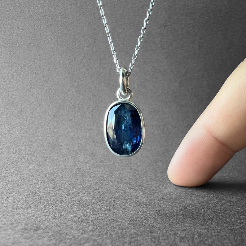May include: A silver pendant necklace featuring an oval-shaped, deep blue gemstone set in a smooth metal bezel. The pendant hangs from a delicate silver chain, showcasing the translucent, faceted quality of the stone with its rich, dark blue hues.