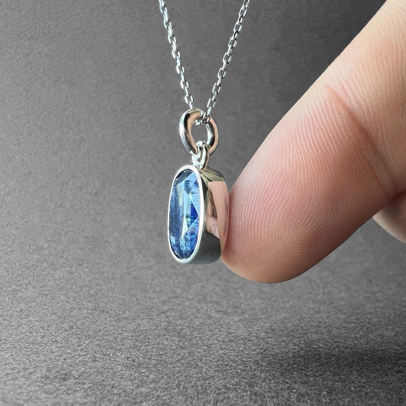 May include: A silver pendant necklace featuring an oval-shaped, faceted blue gemstone set in a polished silver bezel. The pendant hangs from a delicate silver chain, showcasing a deep blue hue with light-reflecting facets throughout the stone.