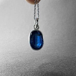 May include: A silver pendant necklace featuring a deep blue, oval-shaped gemstone set in a smooth metal bezel. The pendant hangs from a delicate silver chain, showcasing a polished finish and a vibrant, translucent blue colour within the stone.