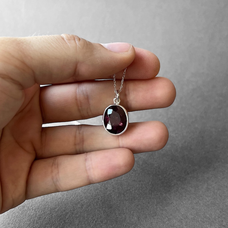 Op de afbeelding: Een ketting met een ovaalvormige, dieppaarse gefacetteerde edelsteen in een gepolijste zilveren zetting. De hanger hangt aan een fijne zilveren ketting en toont een donkere, doorschijnende steen met een glad, reflecterend oppervlak en zilveren rand.