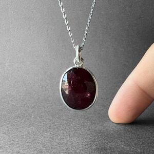 Op de afbeelding: Een zilveren ketting met een ovaalvormige, dieprode gefacetteerde edelsteen hanger. De hanger wordt vastgehouden door een eenvoudig zilveren oogje en hangt aan een fijne zilveren ketting, met een donker, doorschijnend en gepolijst oppervlak.