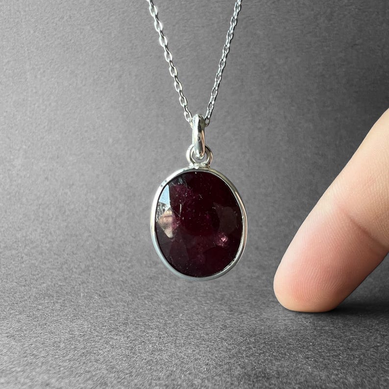 Op de afbeelding: Een zilveren ketting met een ovaalvormige, dieprode gefacetteerde edelsteen hanger. De hanger wordt vastgehouden door een eenvoudig zilveren oogje en hangt aan een fijne zilveren ketting, met een donker, doorschijnend en gepolijst oppervlak.