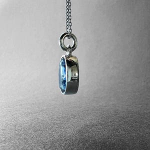 May include: A silver-toned pendant necklace featuring a round, faceted blue gemstone set in a polished metal bezel. The pendant hangs from a delicate silver-toned chain, showcasing a side profile view of the stone's depth and the metal setting.