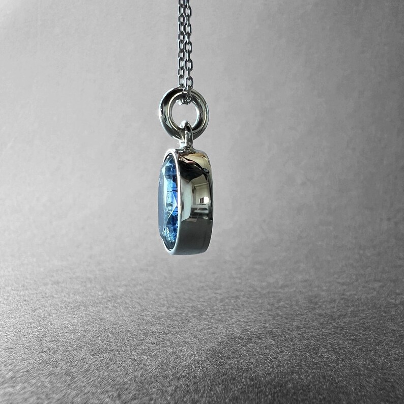 May include: A silver-toned pendant necklace featuring a round, faceted blue gemstone set in a polished metal bezel. The pendant hangs from a delicate silver-toned chain, showcasing a side profile view of the stone's depth and the metal setting.