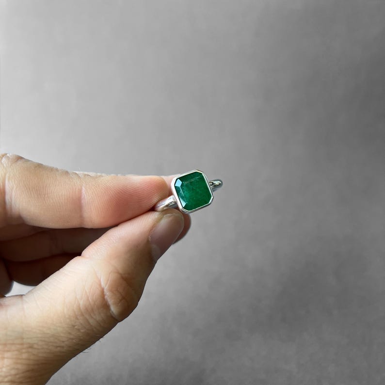K&ouml;nnte beinhalten: Ein Silberring mit einem quadratisch geschliffenen, tiefgr&uuml;nen Edelstein in einer glatten Zargenfassung. Der Ring besteht aus poliertem Silber und der Stein ist mittig platziert, was einen lebendigen, ges&auml;ttigten Gr&uuml;nton und eine facettierte Oberfl&auml;che zeigt.