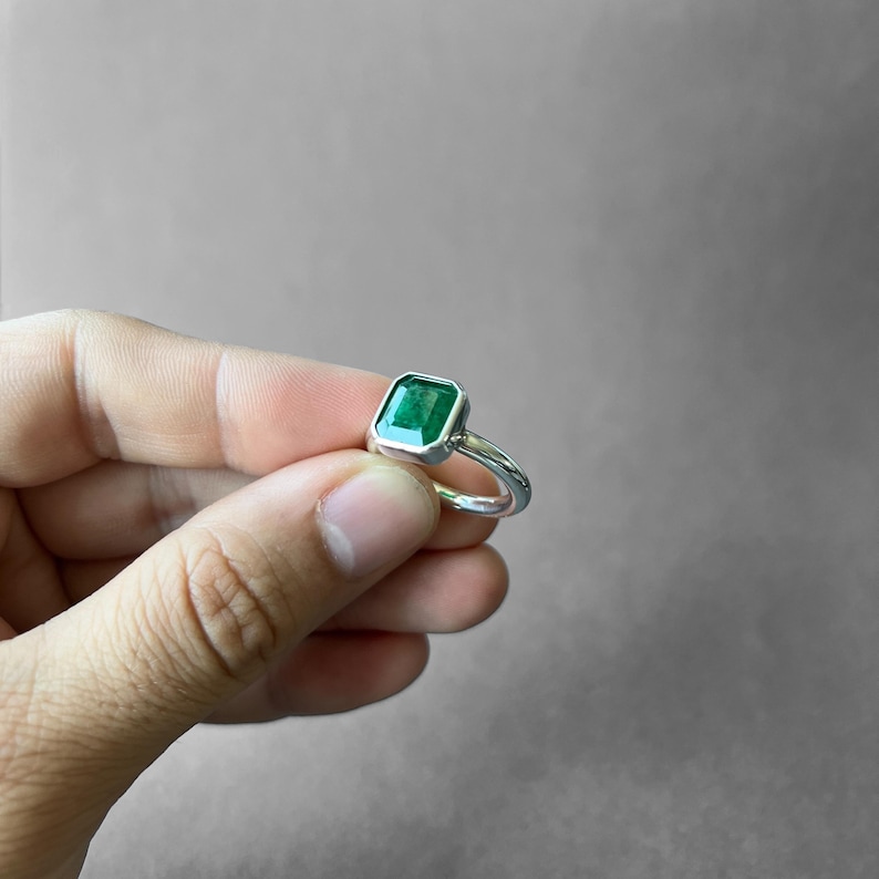 K&ouml;nnte beinhalten: Ein Silberring mit einem rechteckigen, smaragdgeschliffenen gr&uuml;nen Edelstein in einer glatten Zargenfassung. Die Ringschiene besteht aus poliertem Silber mit abgerundetem Profil und pr&auml;sentiert den leuchtend gr&uuml;nen Stein in der Mitte.