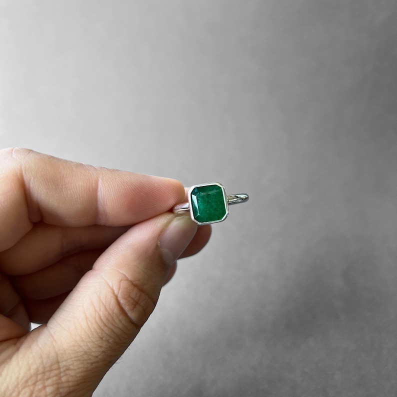 K&ouml;nnte beinhalten: Ein Silberring mit einem rechteckigen, smaragdgeschliffenen gr&uuml;nen Edelstein in einer glatten Zargenfassung. Das Band besteht aus poliertem Silber und der Stein hat eine tiefe, leuchtende gr&uuml;ne Farbe mit einer facettierten Oberfl&auml;che.