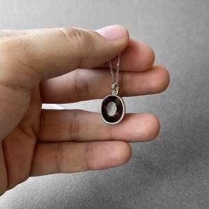 Op de afbeelding: Een zilveren ketting met een ovaalvormige, gefacetteerde dieprode edelsteen hanger in een gladde metalen zetting. De hanger hangt aan een fijne zilveren ketting en heeft een gepolijste afwerking met een diepe, rijke karmozijnrode kleur in de steen.