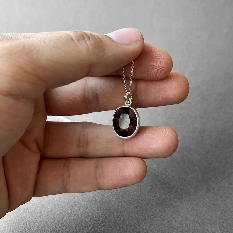 Op de afbeelding: Een zilveren ketting met een ovaalvormige, gefacetteerde dieprode edelsteen hanger in een gladde metalen zetting. De hanger hangt aan een fijne zilveren ketting en heeft een gepolijste afwerking met een diepe, rijke karmozijnrode kleur in de steen.