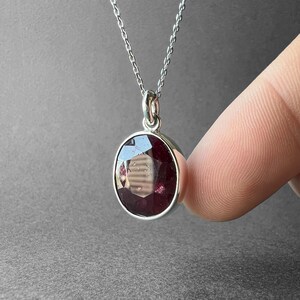 Op de afbeelding: Een zilveren ketting met een ronde, gefacetteerde dieprode edelsteen hanger in een gepolijste zilveren zetting. De hanger hangt aan een fijne zilveren ketting en vertoont een donkerrode kleur met zichtbare facetten en lichtreflecties.