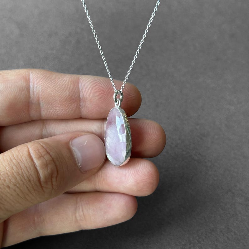 May include: A silver chain necklace with a pink oval pendant. The pendant is made of a translucent stone with a faceted surface.