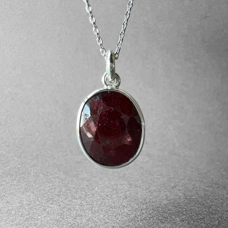 Op de afbeelding: Een zilveren ketting met een ovaalvormige, gefacetteerde donkerrode edelsteen hanger. De steen is gezet in een gladde zilveren rand en hangt aan een fijne zilveren ketting. De hanger heeft een gepolijste afwerking en een diepe rode kleur.