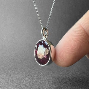 Op de afbeelding: Een zilveren ketting met een ovaalvormige, gefacetteerde donkerrode edelsteen hanger in een gladde metalen zetting. De hanger hangt aan een fijne zilveren ketting en heeft een gepolijste afwerking met een helder, reflecterend oppervlak op de steen.