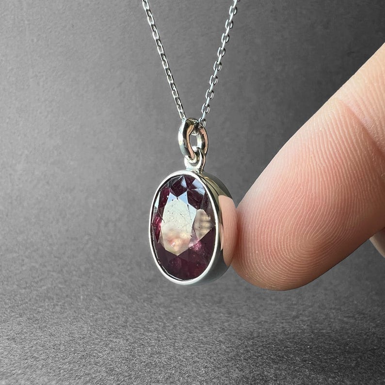 Op de afbeelding: Een zilveren ketting met een ovaalvormige, gefacetteerde donkerrode edelsteen hanger in een gladde metalen zetting. De hanger hangt aan een fijne zilveren ketting en heeft een gepolijste afwerking met een helder, reflecterend oppervlak op de steen.