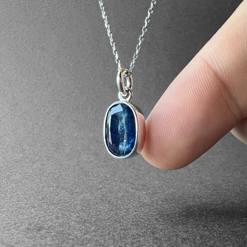 May include: A silver necklace featuring an oval-shaped, faceted blue gemstone pendant. The stone displays deep indigo and lighter blue tones with a translucent quality, held in a simple silver setting attached to a delicate silver chain link.
