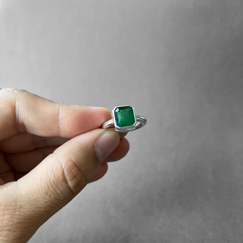 K&ouml;nnte beinhalten: Ein Silberring mit einem rechteckigen, smaragdgeschliffenen gr&uuml;nen Edelstein in einer glatten Metallfassung. Der Stein hat einen tiefen, ges&auml;ttigten gr&uuml;nen Farbton und ist auf einem polierten, abgerundeten Silberring befestigt.