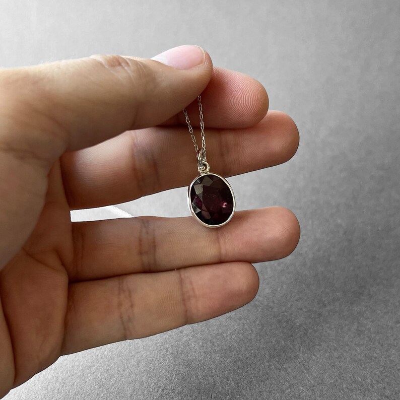 Op de afbeelding: Een zilveren ketting met een ovaalvormige, gefacetteerde dieppaarse edelsteen hanger. De steen is gezet in een eenvoudige zilveren zetting en hangt aan een fijne, dunne zilveren ketting, met een gepolijste en reflecterende oppervlakteafwerking.