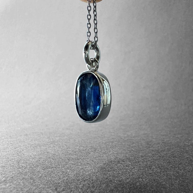 May include: A silver pendant necklace featuring an oval-shaped, deep blue kyanite gemstone set in a polished silver bezel. The pendant hangs from a delicate silver chain, showcasing the stone's natural inclusions and vibrant blue colour tones.