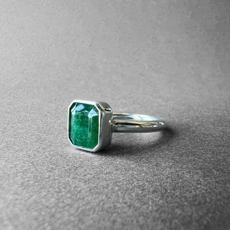 K&ouml;nnte beinhalten: Ein silberner Ring mit einem rechteckigen, smaragdgeschliffenen gr&uuml;nen Edelstein in einer glatten Metallfassung. Die Ringschiene ist poliert und abgerundet und zeigt ein schlichtes Design, das die leuchtend gr&uuml;ne Farbe und die facettierte Oberfl&auml;che betont.