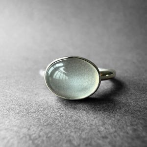 May include: A silver ring featuring a large, oval-shaped, pale blue gemstone. The gemstone is set in a simple, polished silver band. The ring is displayed on a gray surface, with the light reflecting off the stone.