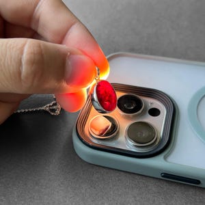 Op de afbeelding: Een ovaalvormige rode edelsteen hanger in een zilveren zetting, verlicht door een lichtbron die een dieprode binnenkant onthult. De hanger zit aan een fijne zilveren ketting en bevindt zich boven de cameralenzen van een smartphone.