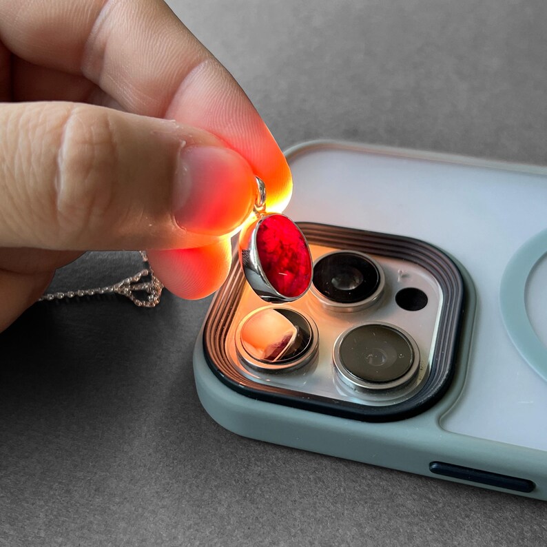 Op de afbeelding: Een ovaalvormige rode edelsteen hanger in een zilveren zetting, verlicht door een lichtbron die een dieprode binnenkant onthult. De hanger zit aan een fijne zilveren ketting en bevindt zich boven de cameralenzen van een smartphone.