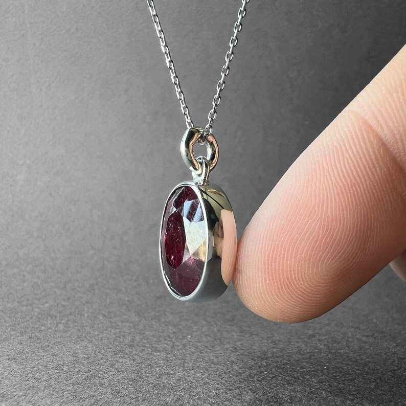 Op de afbeelding: Een zilveren hangerketting met een ovaalvormige, gefacetteerde dieprode edelsteen in een gepolijste zilveren zetting. De hanger hangt aan een fijne zilveren ketting en toont het reflecterende oppervlak en de rijke, donkerrode kleur van de steen.