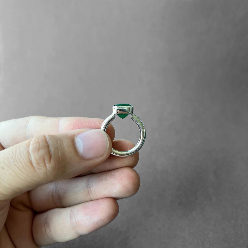 K&ouml;nnte beinhalten: Ein Silberring mit einem einzelnen, facettierten, tiefgr&uuml;nen Edelstein, der in einer glatten, polierten Metallfassung sitzt. Die Ringschiene ist rund und massiv und weist eine saubere, metallische Oberfl&auml;che auf, die das Licht reflektiert.
