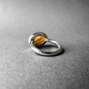 May include: A silver ring with a gold oval-shaped stone set in the center. The ring is shown from the side, with the stone facing the viewer.