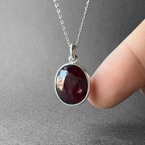 Op de afbeelding: Een zilveren ketting met een ronde, gefacetteerde, donkerrode edelsteen hanger. De steen is gezet in een eenvoudige zilveren zetting en hangt aan een fijne zilveren ketting, met een gepolijste afwerking en reflecterende oppervlakken.