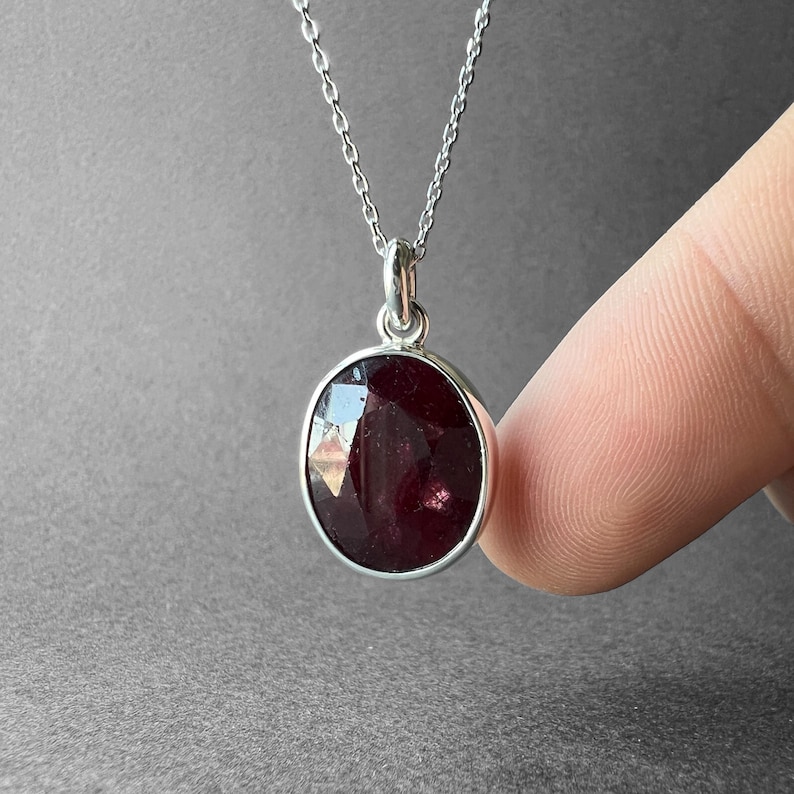 Op de afbeelding: Een zilveren ketting met een ronde, gefacetteerde, donkerrode edelsteen hanger. De steen is gezet in een eenvoudige zilveren zetting en hangt aan een fijne zilveren ketting, met een gepolijste afwerking en reflecterende oppervlakken.